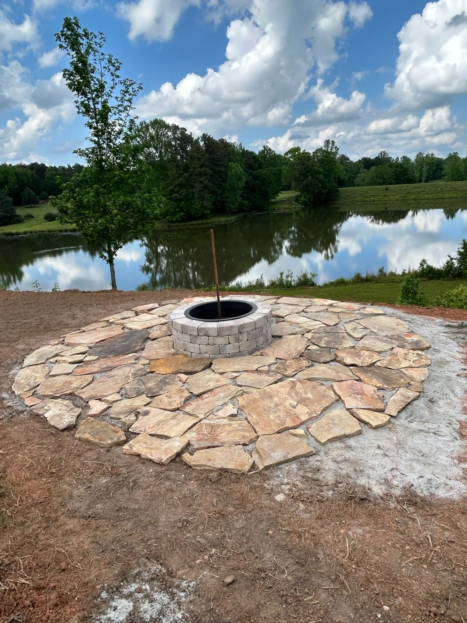 Stone circular fire pit on a stone patio overlooking a calm lake with tree-lined shore under a cloudy sky.