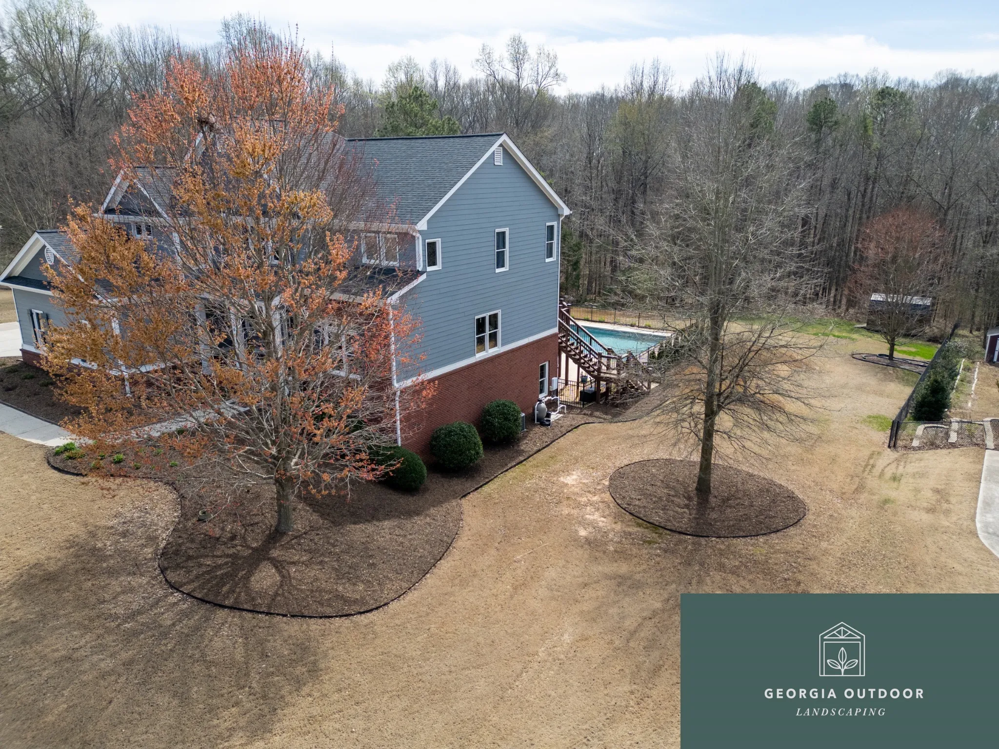 Aerial view of a blue two-story house with a brick base, wooden deck, and a pool, surrounded by bare trees and a wooded background.