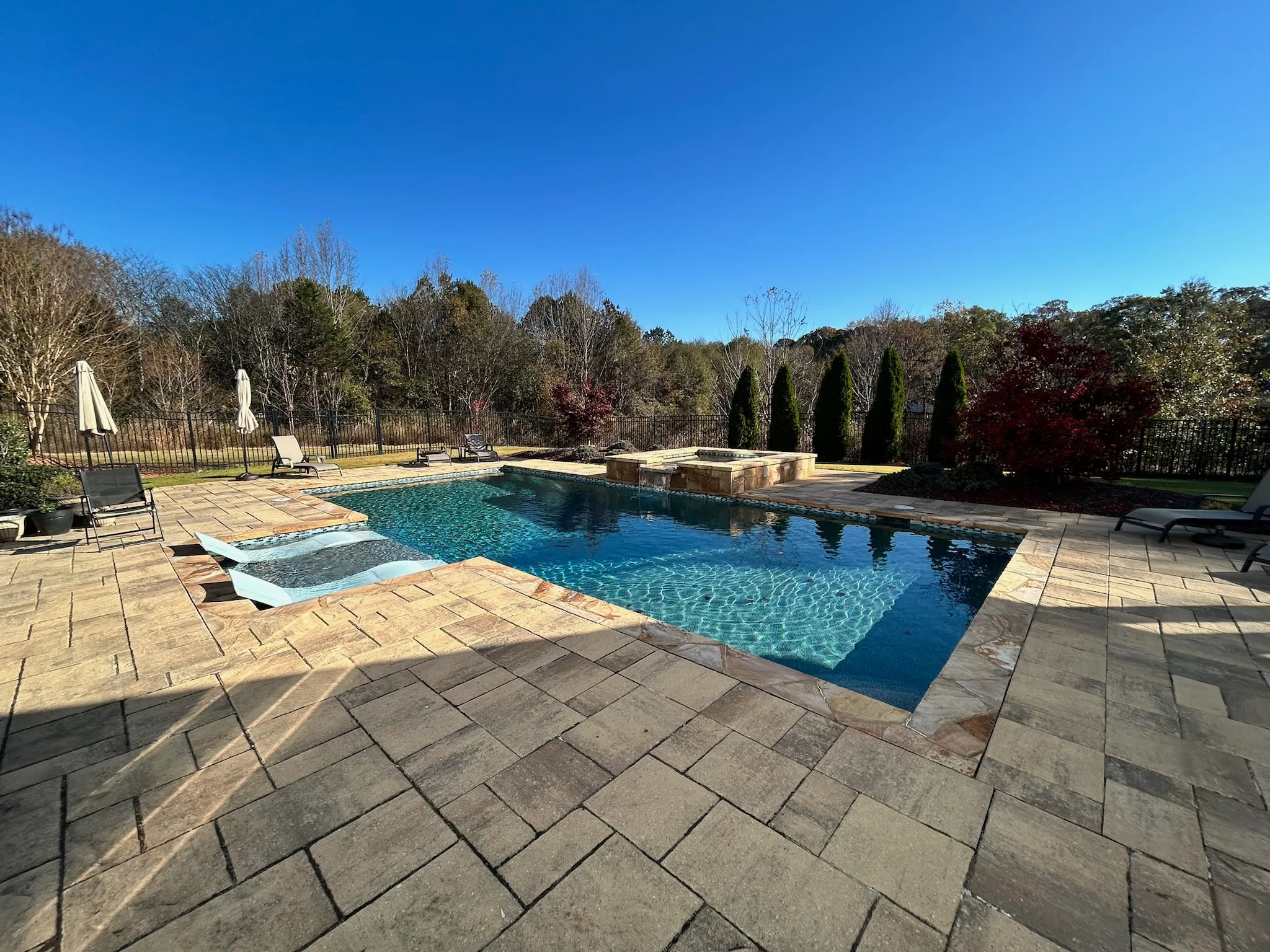 Empty outdoor pool area with beige stone patio, closed umbrellas, and lounge chairs under a clear blue sky, surrounded by trees and a fence.
