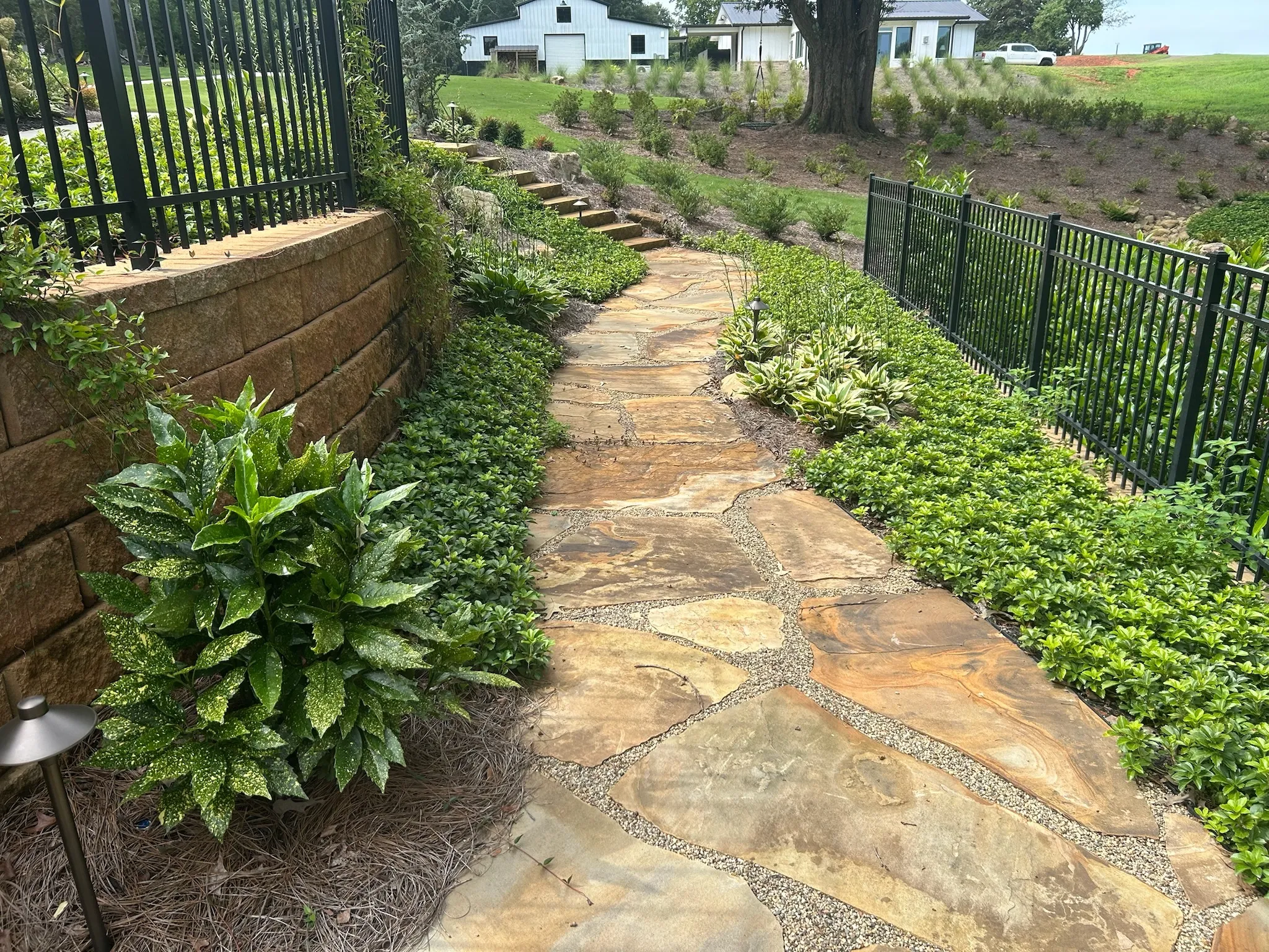 Stone garden path with irregular flagstones, flanked by a stone retaining wall on the left and a black metal fence, surrounded by green shrubs and plants.