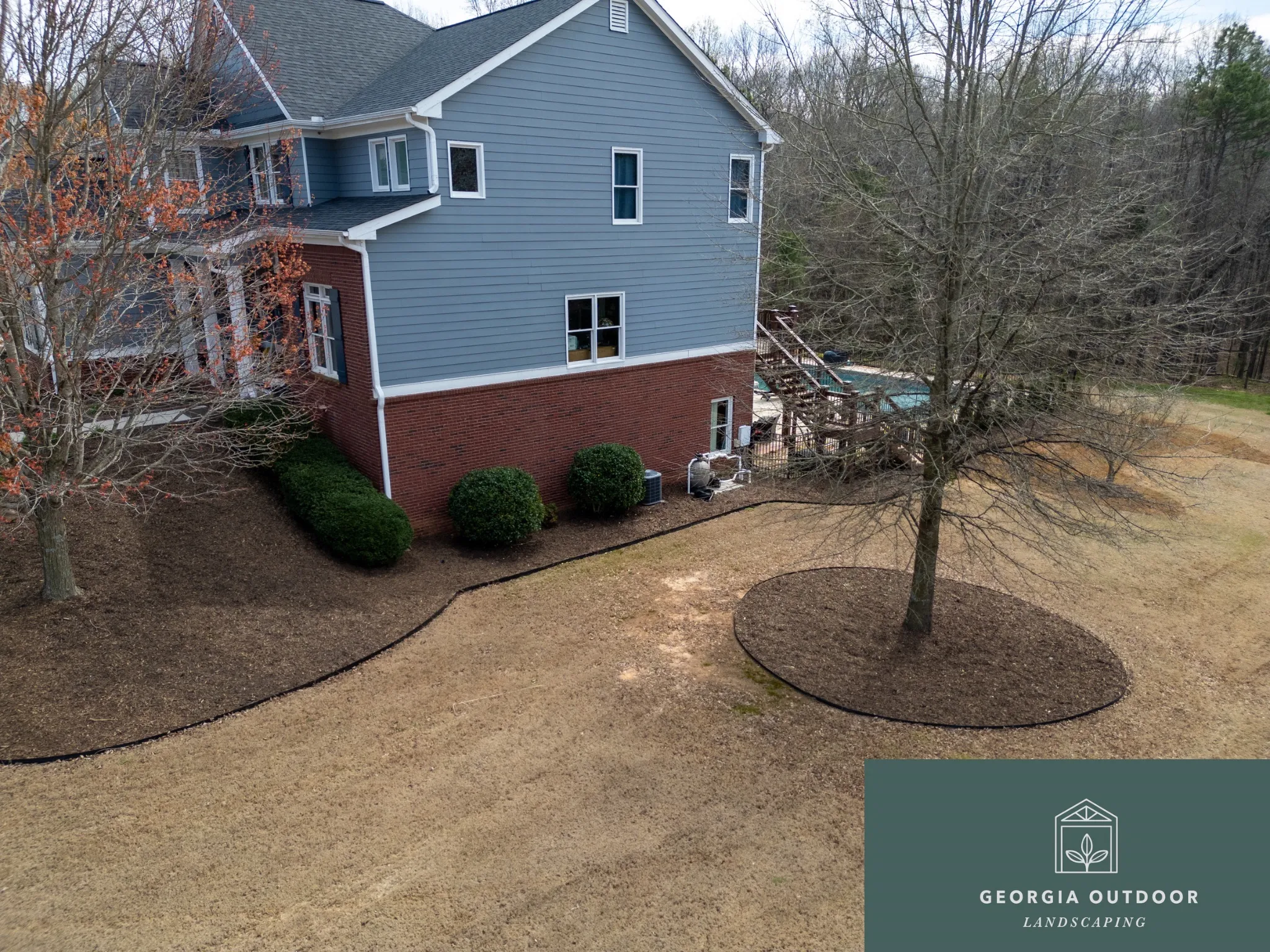 Blue two-story house with brick foundation and white trim, seen from a sloped yard with leafless trees and a wooden deck in the background.