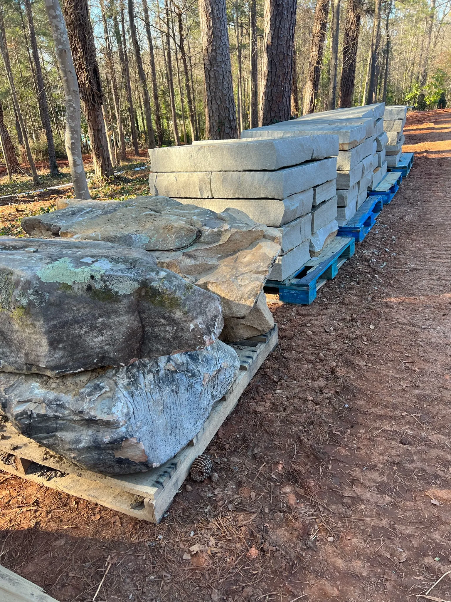 Stacks of light gray rectangular stone blocks on blue pallets in a forested area, ready for construction.
