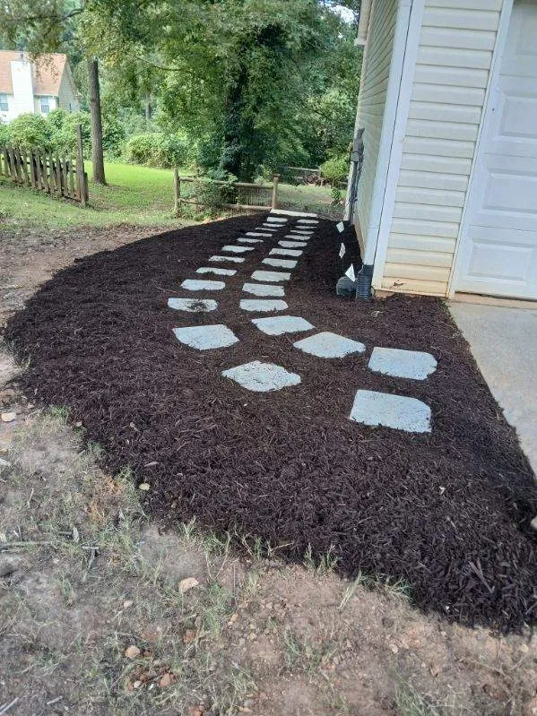 Curved path of white stepping stones set in dark mulch beside a house and garage door leading toward a wooden fence and trees in the yard