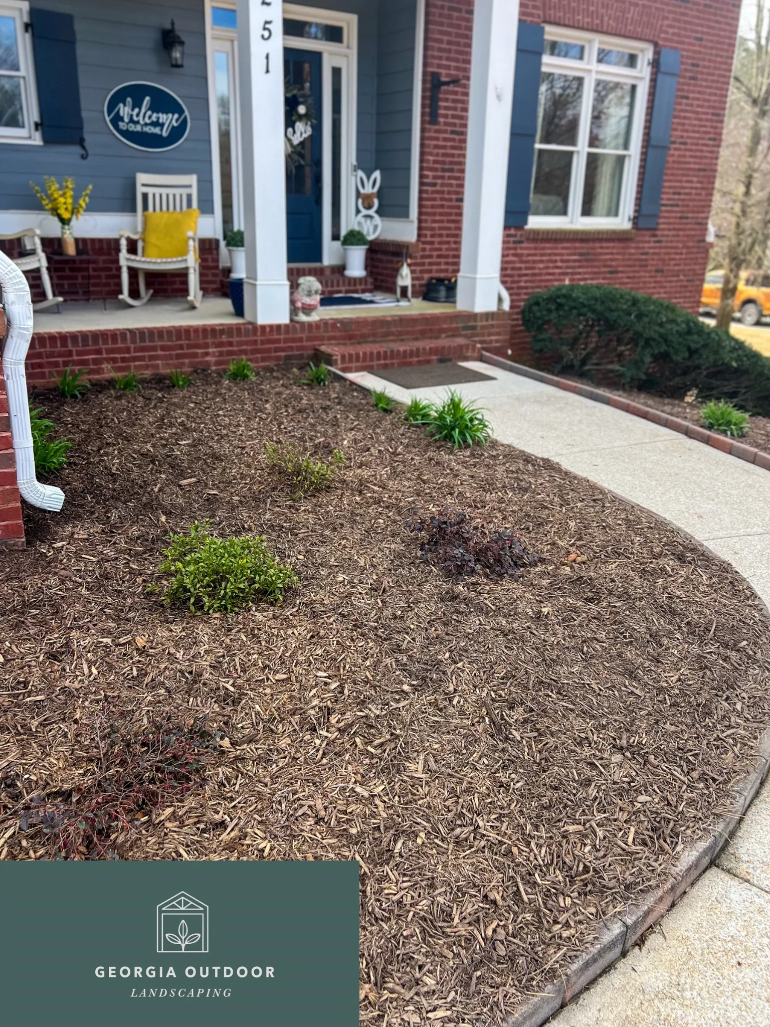 Front porch of a brick house with blue siding, white columns, and a welcome sign; mulched garden beds with small shrubs along a concrete path leading to the porch, and a Georgia Outdoor Landscaping logo overlay in the bottom-left corner.