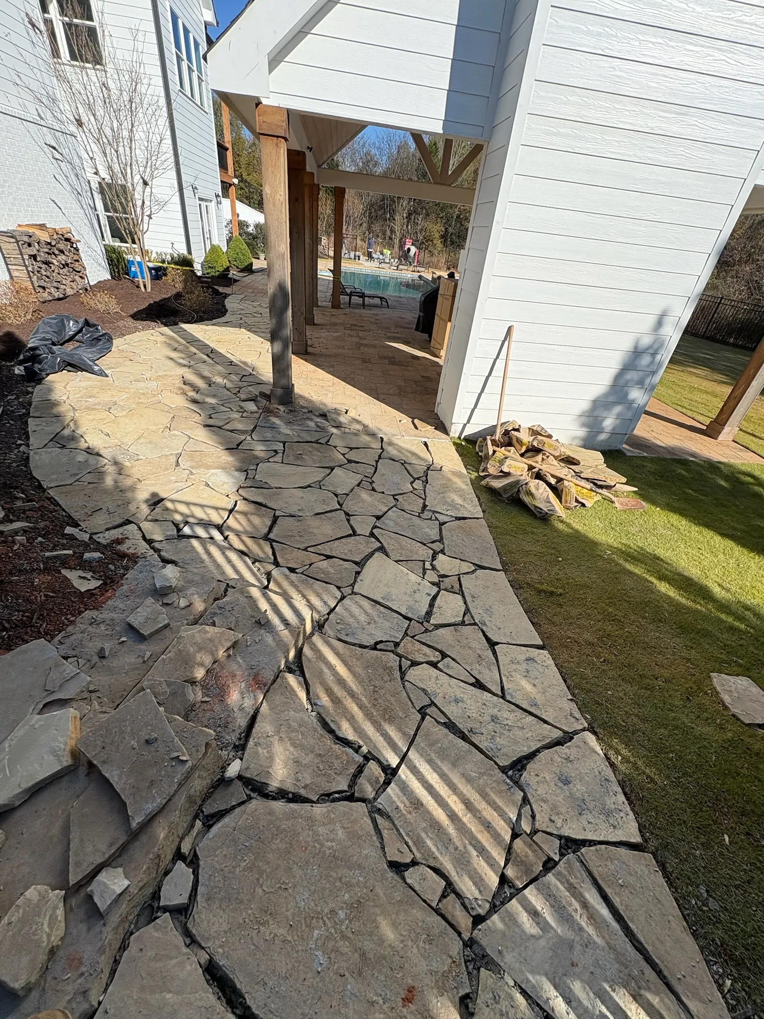 Cracked flagstone patio beside a pool, with wooden support posts and a white house in the background. Bags and construction debris are on the ground.