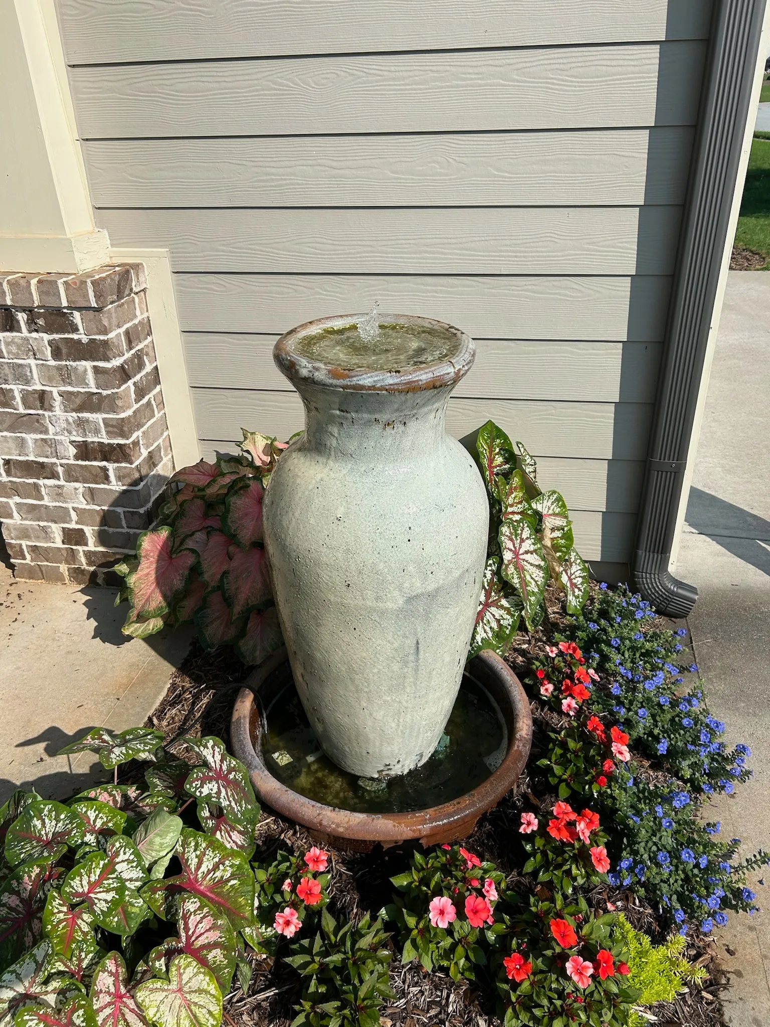 Large weathered ceramic urn fountain with water spouting from the top, surrounded by colorful flowers near the house.