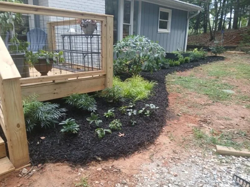 Wooden deck railing with potted plants overlooks a mulched garden bed along a blue house, with new plantings and a gravel path nearby.