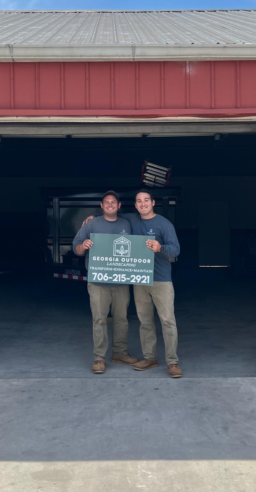 Two men in matching gray shirts stand in a garage holding a Georgia Outdoor Landscaping sign with their phone number: 706-215-2921.